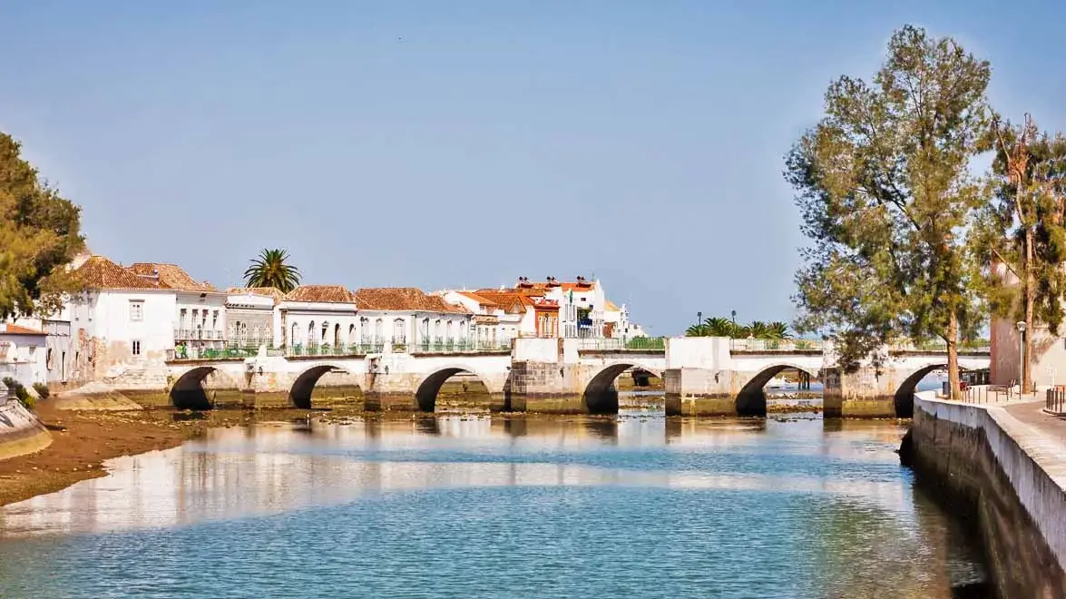 Scenic view of the calm Gilão River in Tavira reflecting the historic old bridge, whitewashed architecture, and palm trees under a bright blue sky
